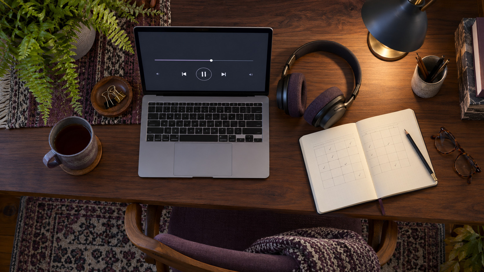 Overhead view of a walnut desk with a laptop audio player, headphones, notebook practice grid and reading glasses.
